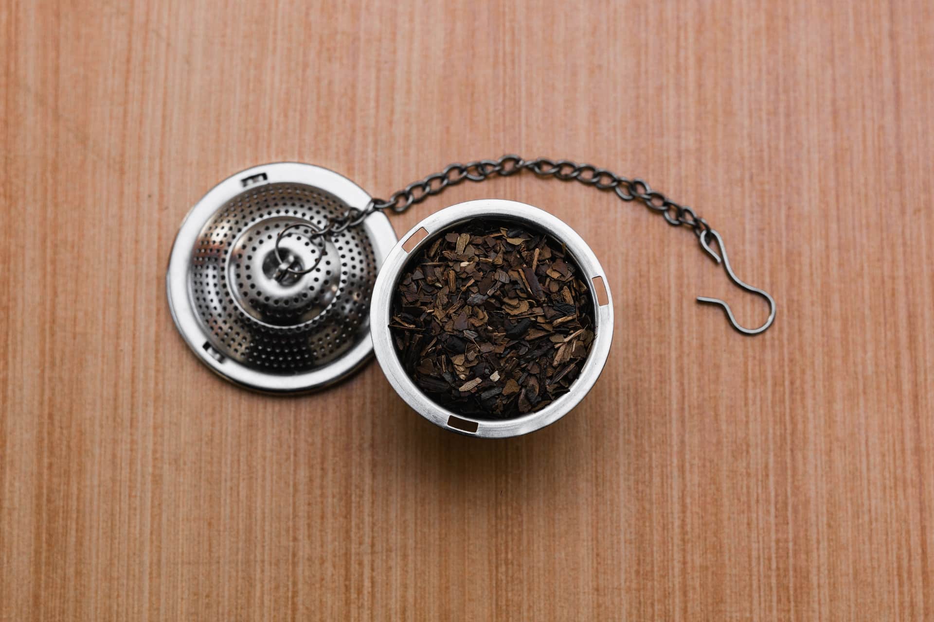 Metal tea infuser filled with loose leaf tea, lying on a wooden surface